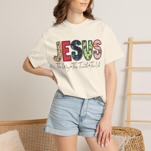 Puede incluir: Una camiseta de manga corta color crema con la palabra JESUS en letras grandes y estampadas. Cada letra muestra un diseño único, incluyendo motivos florales, de cuadros y de rayas. Debajo del texto, la frase The Way The Truth The Life está impresa en una fuente cursiva negra.