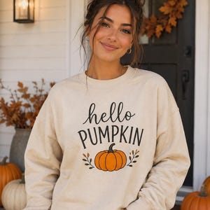 Puede incluir: Una sudadera beige de cuello redondo con el texto negro hello PUMPKIN impreso en la parte delantera. Debajo del texto hay un gráfico de una calabaza naranja con pequeñas hojas marrones a ambos lados. La sudadera tiene mangas largas y cuello.