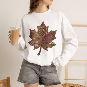 Puede incluir: Una sudadera blanca de manga larga con un gran gráfico central de una hoja de arce. La hoja está dividida en secciones con varios patrones, incluyendo cuadros, flores y estampados de animales en tonos marrones, naranjas y verdes.