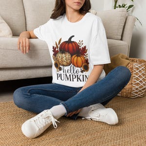 Puede incluir: Una camiseta blanca de manga corta con un estampado gráfico de tres calabazas en tonos naranja y crema, rodeadas de hojas de otoño y girasoles, con el texto negro "hello PUMPKIN" impreso debajo de la ilustración de las calabazas.