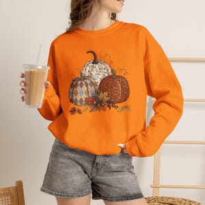 Puede incluir: Una sudadera de color naranja brillante de manga larga con un gráfico central de tres calabazas con distintos patrones, incluyendo un estampado floral, un diseño de rombos azul y beige, y una textura de celosía, decorado con hojas de otoño.