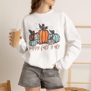 Puede incluir: Una sudadera blanca de manga larga con un gráfico de cuatro calabazas en tonos naranja y azul verdoso, con el texto "happy fall y'all" impreso en cursiva marrón debajo. La sudadera tiene cuello redondo y puños acanalados.