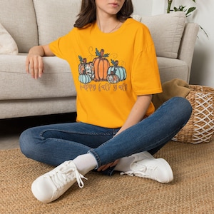 May include: A bright yellow short-sleeve t-shirt featuring a graphic of four pumpkins in shades of orange, teal, and patterned brown. Below the pumpkins, the text reads "happy fall y'all" in a cursive brown font. The shirt has a crew neckline.