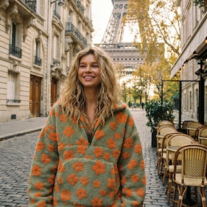 Könnte beinhalten: Ein langärmeliger Pullover mit kurzem Reißverschluss aus flauschigem, olivgrünem Fleece-Material mit einem sich wiederholenden Muster aus leuchtend orangefarbenen Blumen. Das Kleidungsstück hat einen hohen Kragen, lange Ärmel und eine große Kängurutasche.