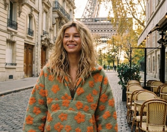 May include: A long-sleeve, quarter-zip pullover made of fuzzy, olive green fleece material featuring a repeating pattern of bright orange flowers. The garment includes a high collar, long sleeves, and a large front kangaroo pocket.
