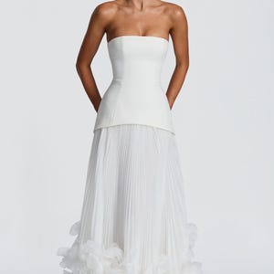 May include: A floor-length white strapless gown featuring a structured bodice with a straight neckline and a dropped waist. The skirt is composed of fine vertical pleats and is finished at the hem with a dense layer of layered white fabric petals.
