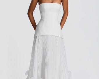 May include: A floor-length white strapless gown featuring a structured bodice with a straight neckline and a dropped waist. The skirt is composed of fine vertical pleats and is finished at the hem with a dense layer of layered white fabric petals.