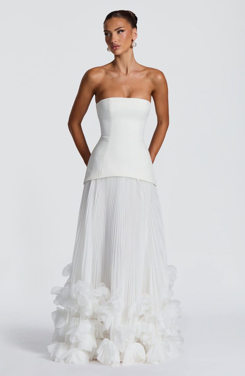 May include: A floor-length white strapless gown featuring a structured bodice with a straight neckline and a dropped waist. The skirt is composed of fine vertical pleats and is finished at the hem with a dense layer of layered white fabric petals.
