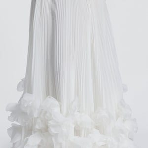 May include: A floor-length white skirt featuring a smooth waistband and a pleated main body. The hem is decorated with a dense layer of layered, semi-sheer white fabric petals that create a voluminous, textured appearance at the bottom edge.