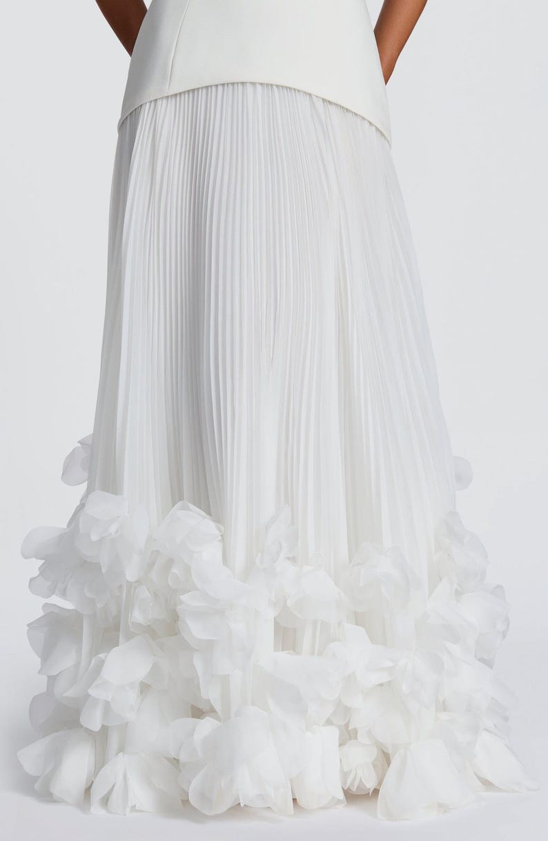 May include: A floor-length white skirt featuring a smooth waistband and a pleated main body. The hem is decorated with a dense layer of layered, semi-sheer white fabric petals that create a voluminous, textured appearance at the bottom edge.