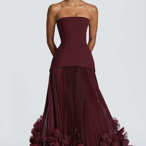 May include: A floor-length strapless gown in a deep burgundy hue. The bodice is structured and fitted, transitioning into a full-length skirt featuring fine vertical pleats and a hemline adorned with layered, ruffled fabric floral accents.