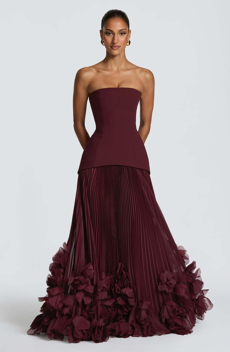May include: A floor-length strapless gown in a deep burgundy hue. The bodice is structured and fitted, transitioning into a full-length skirt featuring fine vertical pleats and a hemline adorned with layered, ruffled fabric floral accents.