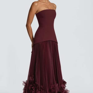 May include: A floor-length strapless gown in a deep burgundy hue. The bodice is fitted and smooth, transitioning into a floor-length skirt featuring fine vertical pleats. The hem is adorned with a dense layer of layered fabric floral appliques.