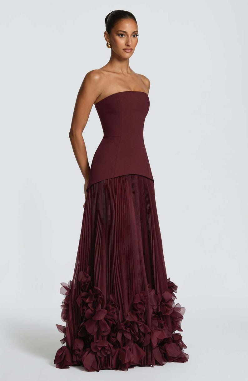 May include: A floor-length strapless gown in a deep burgundy hue. The bodice is fitted and smooth, transitioning into a floor-length skirt featuring fine vertical pleats. The hem is adorned with a dense layer of layered fabric floral appliques.