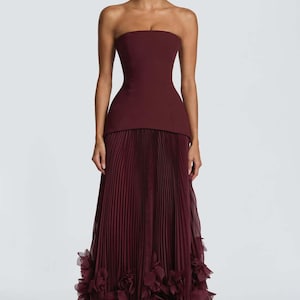 May include: A floor-length strapless gown in a deep burgundy hue. The bodice is fitted and structured, transitioning into a long, pleated skirt. The hemline is adorned with voluminous, layered fabric ruffles that create a textured, floral-like effect.