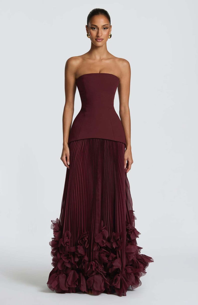 May include: A floor-length strapless gown in a deep burgundy hue. The bodice is fitted and structured, transitioning into a long, pleated skirt. The hemline is adorned with voluminous, layered fabric ruffles that create a textured, floral-like effect.