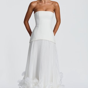 May include: A floor-length white strapless gown featuring a structured bodice with a straight neckline and a long, pleated skirt. The hem of the skirt is adorned with a dense layer of white fabric petals, creating a voluminous, textured finish.
