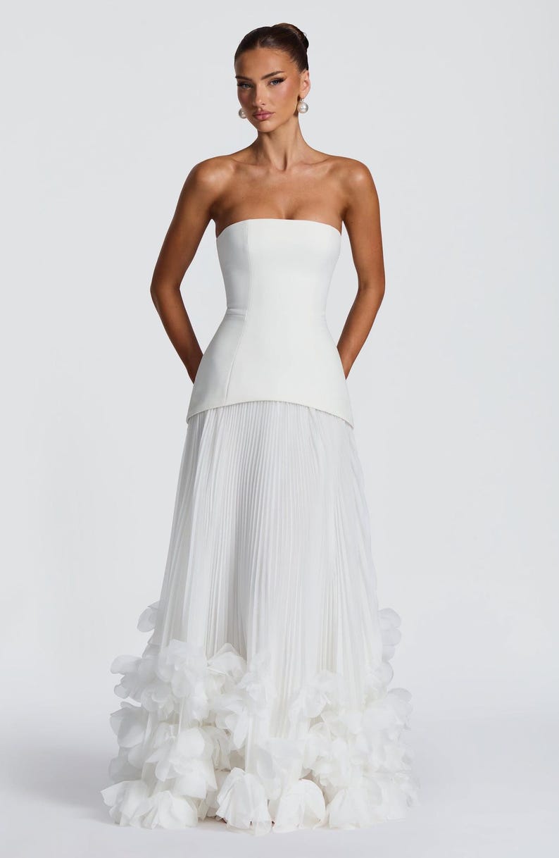May include: A floor-length white strapless gown featuring a structured bodice with a straight neckline and a long, pleated skirt. The hem of the skirt is adorned with a dense layer of white fabric petals, creating a voluminous, textured finish.
