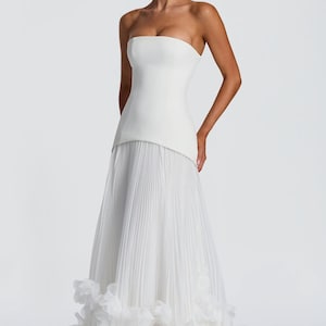 May include: A floor-length white strapless gown featuring a structured bodice and a pleated skirt. The hem of the skirt is adorned with layered, ruffled fabric accents that create a voluminous texture at the base of the garment.