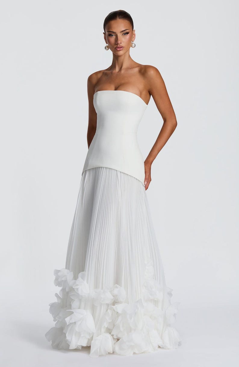 May include: A floor-length white strapless gown featuring a structured bodice and a pleated skirt. The hem of the skirt is adorned with layered, ruffled fabric accents that create a voluminous texture at the base of the garment.