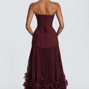 May include: A floor-length burgundy gown featuring a strapless corset-style bodice with a lace-up back closure. The skirt is constructed with fine vertical pleats and finished with a voluminous hemline adorned with layered fabric floral rosettes.