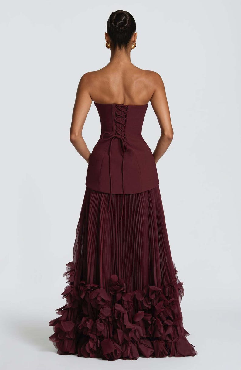 May include: A floor-length burgundy gown featuring a strapless corset-style bodice with a lace-up back closure. The skirt is constructed with fine vertical pleats and finished with a voluminous hemline adorned with layered fabric floral rosettes.
