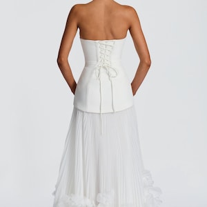 May include: A white strapless gown featuring a structured corset-style bodice with a lace-up back closure. The skirt is composed of fine vertical pleats that extend to the floor, finished with a layered, ruffled hemline at the bottom edge.