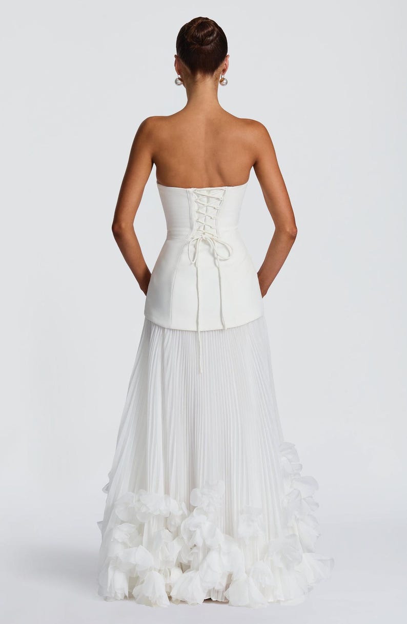 May include: A white strapless gown featuring a structured corset-style bodice with a lace-up back closure. The skirt is composed of fine vertical pleats that extend to the floor, finished with a layered, ruffled hemline at the bottom edge.