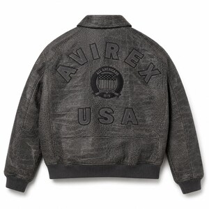 Op de afbeelding: Een donkergrijze jas met een textuur van krokodillenleer. De achterkant is voorzien van de tekst AVIREX in een boog, een ronde patch met de tekst ALL AMERICANS ESTABLISHED 1975 en een schildafbeelding, en de tekst USA in grote letters.