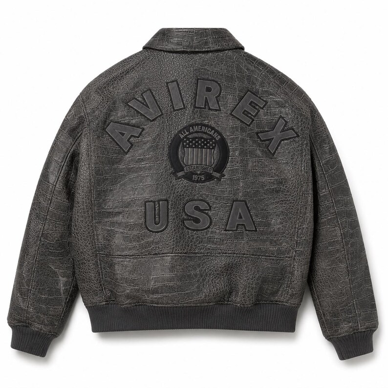 Op de afbeelding: Een donkergrijze jas met een textuur van krokodillenleer. De achterkant is voorzien van de tekst AVIREX in een boog, een ronde patch met de tekst ALL AMERICANS ESTABLISHED 1975 en een schildafbeelding, en de tekst USA in grote letters.