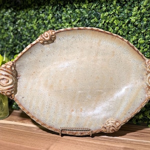May include: An oval ceramic serving platter with a speckled beige and cream glaze. The platter features a raised, textured brown rim and four decorative, spiral-shaped handles or accents positioned symmetrically around the edge of the dish.