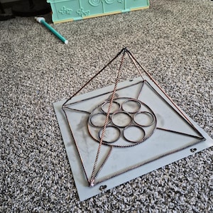 Op de afbeelding: Een koperkleurige metalen piramidestructuur met een vierkante bodemplaat en een centraal cirkelvormig patroon van zeven onderling verbonden ringen. Het piramideframe is opgebouwd uit gedraaide metalen staven.