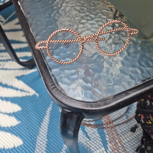 May include: A pair of eyeglasses crafted from twisted copper wire, featuring two circular frames connected by a central bridge. The wire has a distinct braided texture throughout the entire structure, creating a metallic, reddish-brown appearance.