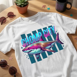 Può includere: Una t-shirt bianca a maniche corte con una stampa grafica di uno squalo nei toni del rosa, blu e viola. Il testo CHOMP CITY è stampato in un carattere blu audace e stilizzato con un contorno rosa sopra lo squalo, su strisce verticali.