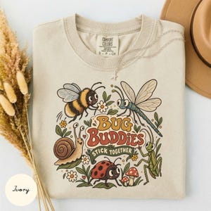 Op de afbeelding: Een ivoorkleurig t-shirt met een kleurrijke afbeelding van een bij, libel, slak, lieveheersbeestje en sprinkhaan omringd door bloemen en paddenstoelen. De tekst luidt Bug Buddies Stick Together in een retro lettertype. Het shirt heeft een ronde hals.