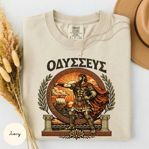 May include: An ivory t-shirt featuring a graphic print of a Greek warrior in armour and a red cape, standing before a sunburst and a ship. The Greek text "ΟΔΥΣΣΕΥΣ" is printed above the circular design, which is framed by laurel leaves and columns.