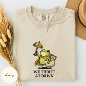 May include: An ivory t-shirt featuring a graphic of a green frog wearing a yellow cape, holding a stained glass lamp and a tote bag containing a framed picture. The text below the frog reads WE THRIFT AT DAWN with small star icons on each side.