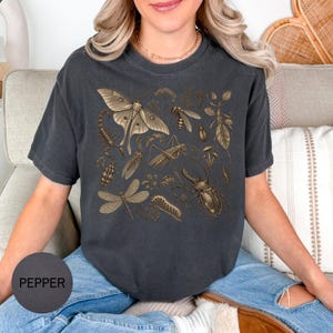 Może przedstawiać: Ciemnoszary t-shirt z krótkim rękawem z detalicznym złotym nadrukiem graficznym przedstawiającym różne owady, w tym ćmę, skorpiona, ważkę, chrząszcza i konika polnego, z okrągłą próbką koloru z napisem PEPPER w rogu.