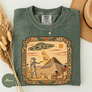 May include: A moss green t-shirt featuring a graphic print of an alien hovering a UFO over an Egyptian pyramid construction site. The design includes a decorative border with bird motifs and the Comfort Colors brand label at the inner neckline.