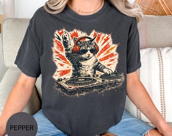 May include: A charcoal gray t-shirt featuring a graphic of a cat wearing red headphones and black sunglasses, with one paw raised while operating a DJ turntable. The background of the graphic consists of orange and red radiating lightning bolts.