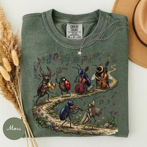 Könnte beinhalten: Ein moosgrünes T-Shirt mit einer Grafik von Insekten, die auf einem Pfad Musikinstrumente spielen, darunter ein Käfer mit Becken, ein Marienkäfer, ein Cello spielender Käfer, eine Biene, ein Käfer mit Geige und eine Heuschrecke mit Akkordeon.