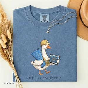 Może przedstawiać: Niebieski t-shirt z grafiką przedstawiającą białą gęś w żółtym kasku budowlanym i beżowej kamizelce. Gęś trzyma niebieski projekt oraz tablet z rysunkiem technicznym. Pod grafiką znajduje się napis "OFF TO ENGINEER".