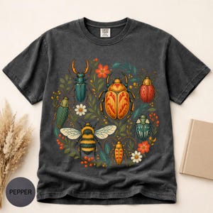 以下が含まれることがあります： チャコールグレーの半袖Tシャツ。中央には色とりどりの甲虫、マルハナバチ、小さな花のグラフィックプリントが施されています。クルーネックで、ヴィンテージ風のウォッシュ加工が施された生地感のTシャツで、Comfort Colorsのラベルが付いています。