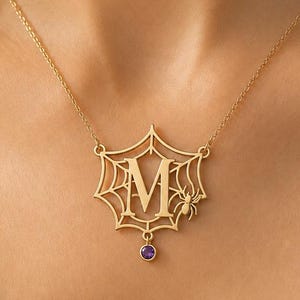 Può includere: Una collana color oro con un ciondolo a forma di ragnatela che presenta la lettera M al centro. Un piccolo ragno è attaccato sul lato destro della ragnatela e una singola pietra preziosa rotonda viola pende dalla parte inferiore del disegno.