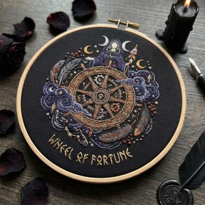 May include: A circular embroidery hoop containing black fabric with a detailed thread-stitched design of a wooden wheel, a hooded figure, feathers, and celestial motifs. The text "WHEEL OF FORTUNE" is embroidered in gold at the bottom.