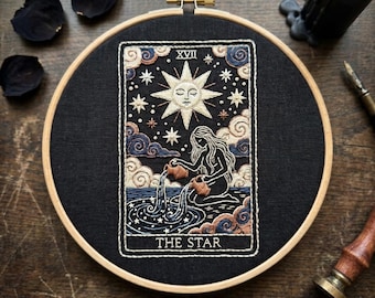May include: A circular wooden embroidery hoop framing a black fabric panel featuring a detailed needlework design of The Star tarot card. The embroidery depicts a figure pouring water from two jugs into a pool beneath a large sun and stars.