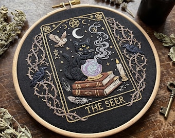 May include: A circular embroidery hoop containing black fabric with a detailed needlework design of a black cat sitting on two brown books, gazing into a crystal ball. The scene includes a crescent moon, an owl, two ravens, and the text THE SEER.