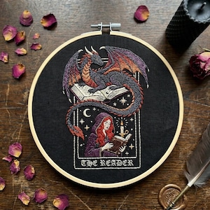 May include: A circular embroidery hoop containing black fabric with a detailed thread-stitched design. The artwork features a dragon perched on an open book above a hooded figure reading by candlelight. The text THE READER is stitched below.