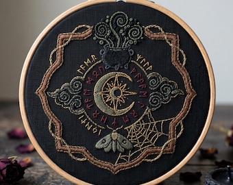 May include: A circular wooden embroidery hoop containing black fabric with intricate needlework. The design features a central crescent moon and star, a cauldron with swirling steam, a moth, a spiderweb, and runic symbols in gold and red thread.