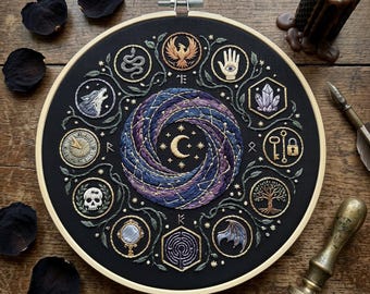 May include: A circular embroidery hoop featuring black fabric with a central swirling galaxy design, a crescent moon, and stars. Surrounding the center are ten embroidered icons including a phoenix, hand, crystal, key, lock, tree, wing, labyrinth, mirror, skull, sundial, wolf, and snake.